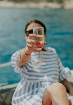 Portrait of one young Caucasian beautiful girl in sunglasses sitting in a boat holding out a glass of champagne and enjoying sailing on the sea on a private boat on a sunny summer day, close-up side view with selective focus.