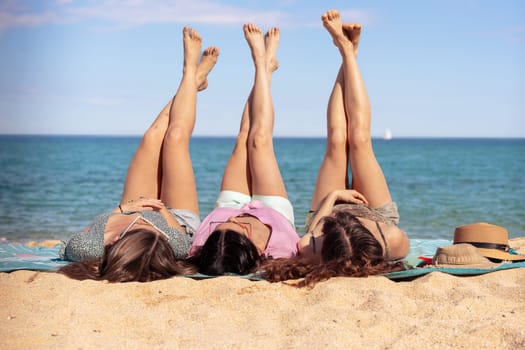 Three young women lying on a tropical beach, stretching their slender legs. Blue sea in the background. Summer vacation concept.