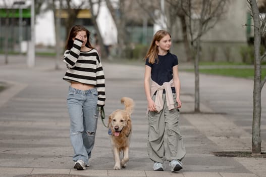 Two Sisters With A Golden Retriever Are Walking On The Street In Summer