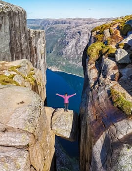 A person stands on a narrow rock formation overlooking a breathtaking fjord in Norway. The cliffs on either side rise high, with deep blue water visible below. Kjeragbolten, Norway