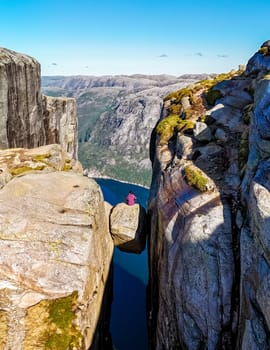 A lone figure sits on the edge of Kjeragbolten, a famous cliff in Norway, with a breathtaking view of the surrounding landscape.