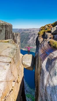 A woman stands on the edge of Kjeragbolten, Norway, a famous cliff in Norway. The view from the top is breathtaking, with a deep valley and a blue fjord in the background.