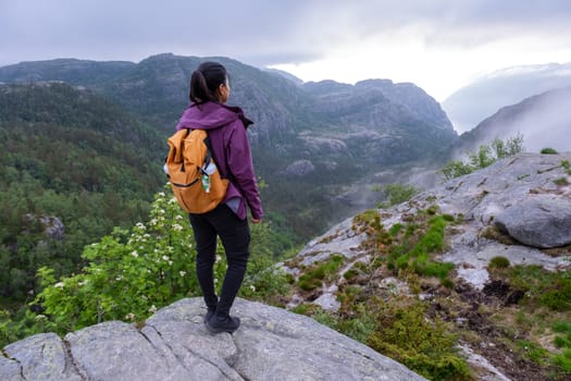 A woman with a backpack stands on a cliffside, gazing at a stunning view of a Norwegian fjord. The mountains are covered in mist, and the landscape is breathtaking. Preikestolen, Norway