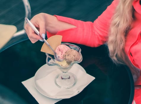 Hands of a young Caucasian unrecognizable woman in a pink shirt with delicate manicure eating chocolate-strawberry ice cream with a waffle while sitting at a black glass table in a street cafe, top side close-up view. Concept of summer food, restaurant ice cream.