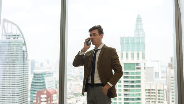 Businessman standing in ornamented office talking on phone with college on cityscape skyline window background. Determination and ambition drive business career toward to bright future