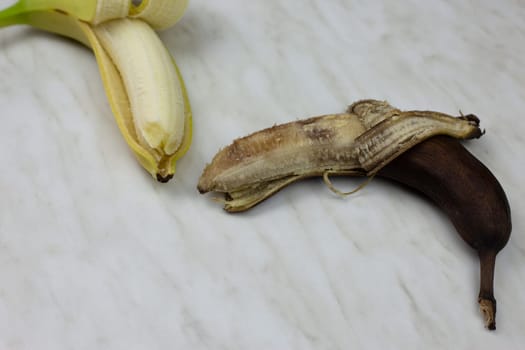 Rotten spoiled banana in open peel next to fresh banana fruit on white table, phase of spoiled fruit