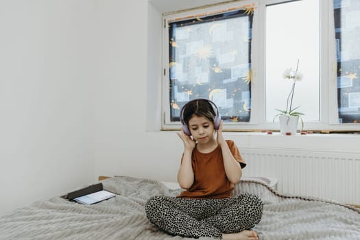 Portrait of a beautiful Caucasian brunette girl with big eyes sitting on a bed in a room by the window listening to her favorite music through headphones on a spring day, view from the side.