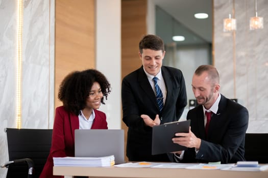 Diverse business team of three people, including a woman and two men, collaborating at an office desk with a laptop and paperwork, bright modern office setting