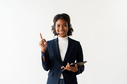 A professional female call center employee wearing a headset, smiling, and using a tablet in a modern office environment.