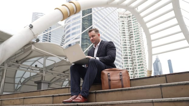 Low angle view of professional business man working on laptop at stairs. Caucasian project manager using computer to plan marketing strategy and communicate with financial team at urban city. Urbane.