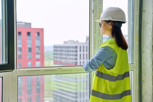 Back view of female industrial worker in protective helmet vest looking at new buildings through window. Construction engineering, real estate sales buying office apartment, work technical professions
