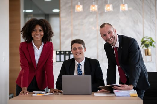 Diverse business team of three people, including a woman and two men, smiling at an office desk with a laptop and paperwork, bright modern office setting