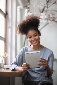 A young woman sits in a bright living room, taking notes in a notebook, smiling and engaged in her work. The room features minimalist decor and natural light.