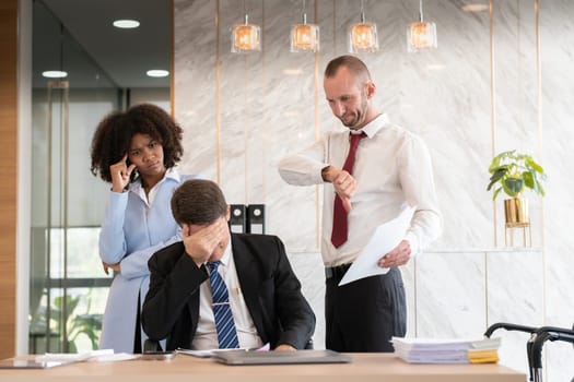 Frustrated diverse business team of three people, including a woman and two men, dealing with paperwork at an office desk, showing stress and disappointment, modern office setting