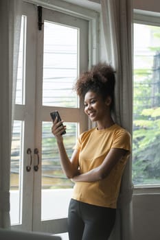 A young African woman smiling while using her smartphone by the window. She is dressed casually and enjoying a relaxed moment at home.