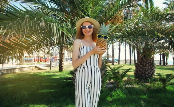 Summer fashion outfit of stylish happy smiling young woman holding pineapple fruits wearing hat, striped clothes, sunglasses posing in park with palm trees background