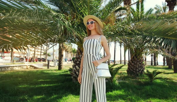 Summer outfit, stylish beautiful young woman posing in white striped jumpsuit with handbag standing in green park with palm trees background