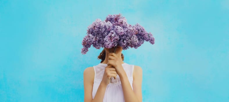 Portrait of woman covering her head with bouquet of fresh lilac flowers on blue background