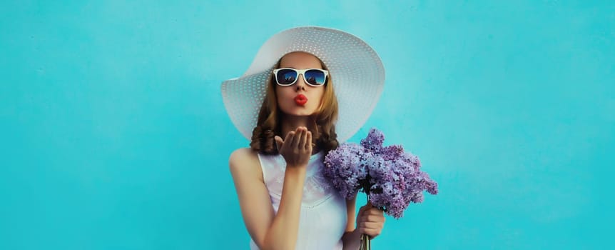 Summer portrait of beautiful lovely young woman blowing kiss with bouquet of purple lilac flowers in white straw hat on blue background