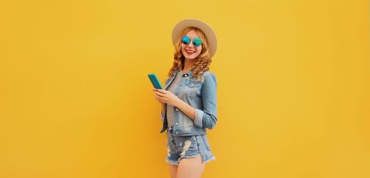 Summer modern happy young woman holding a smartphone in straw tourist hat, denim clothing posing on yellow wall background