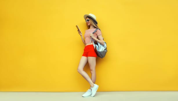 Full-length summer vacation, tourism, happy traveler tourist young woman holding smartphone looking at device in casual straw hat with backpack on bright yellow background