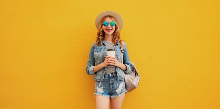 Beautiful stylish happy smiling young woman standing with cup of coffee in summer hat, backpack, jean jacket on yellow background