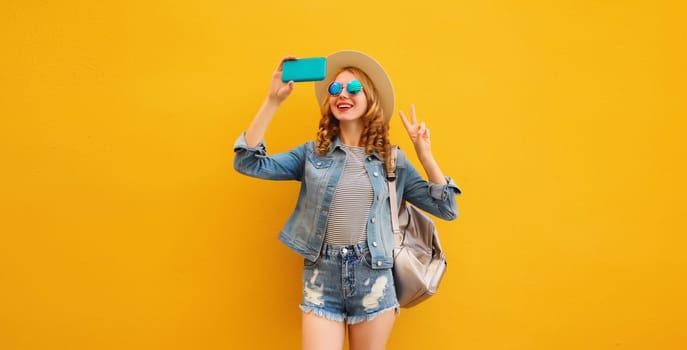 Portrait of stylish happy smiling young woman taking selfie with phone and backpack in summer tourist straw hat, denim clothing on bright yellow background
