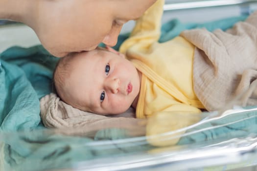 Mom kisses her newborn baby who is resting in a transparent cradle in the hospital. This tender moment emphasizes the bond between mother and child, creating a cozy and safe atmosphere for the infant