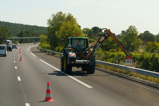 Salles-d'Aude, France - May 16, 2023: A maintenance worker operates an excavator on the side of a highway, with traffic cones set up for safety during the daytime.