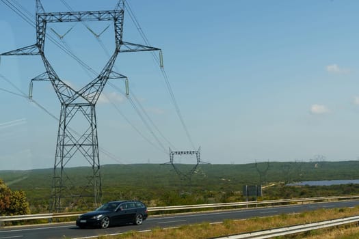 A black car drives on a highway, passing under a large power line tower in a rural landscape. The sky is blue and clear.