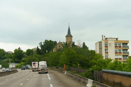 Orange, France - May 16, 2023: A view of a highway in France with trucks driving past a church and an apartment building.