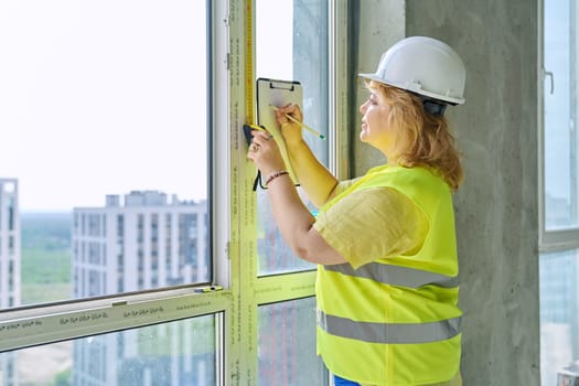 Female window decorator designer measuring window with tape measure, writing down measurements for sewing curtains making blinds in new apartment office. Decorating design service