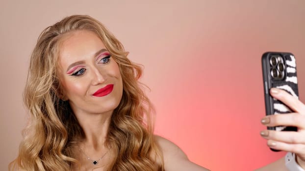 A young woman with red lipstick takes a selfie with long blond hair and makeup. The girl is smiling and looking at the camera close-up