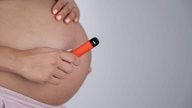 A pregnant woman smokes a vape. A girl holds an electronic cigarette against the background of her bare tummy. Copy space