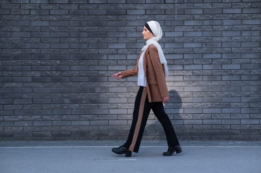 A young woman dressed in a hijab and a business suit walks along a brick wall