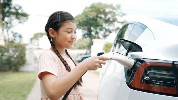 Happy little young girl learn about eco-friendly and energy sustainability as she recharge electric vehicle from home EV charging station. EV car and sustainable future generation concept. Synchronos