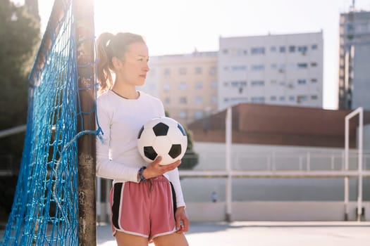 caucasian female football player holding a soccer ball leaning on the goal of an urban court, concept of sport and active lifestyle, copy space for text
