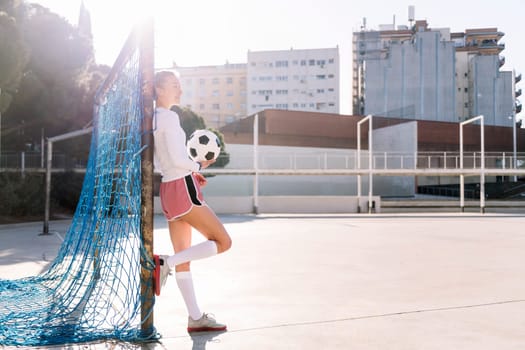 smiling caucasian female football player holding a soccer ball leaning on the goal of an urban court, concept of sport and active lifestyle, copy space for text