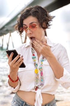 Caucasian woman wearing sunglasses vacationing by the sea with a gesture of surprise using a social media app with her mobile phone