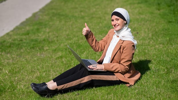 A young woman wearing a hijab sits on a lawn and uses a laptop outdoors and shows a thumbs up