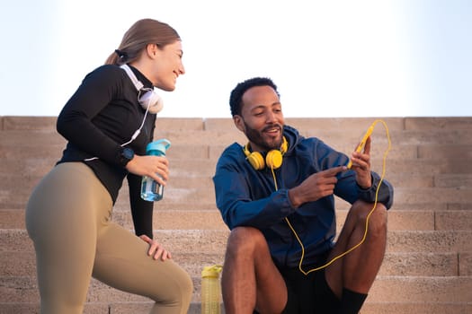 Smiling man using a social media app on a smartphone and young woman drinking water in sports bottle while exercising outdoors.