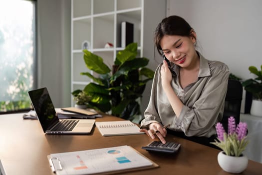 Young woman working from home office, using a calculator and smiling, surrounded by modern office supplies and indoor plants in a bright, minimalist workspace.