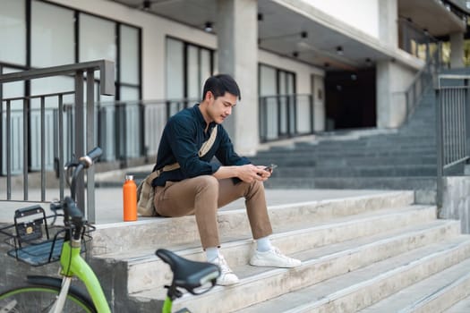 A young professional using a smartphone while sitting on steps next to a green bicycle, embodying a business eco lifestyle in an urban environment.