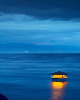 Twilight over a calm sea with a solitary, illuminated floating hut and distant city lights.