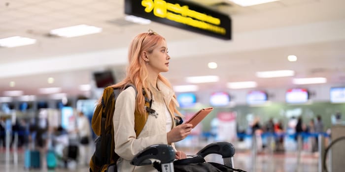 A young woman with luggage and a tablet stands in a modern airport terminal, ready for her journey. The image captures the essence of travel and technology in a busy, contemporary setting.