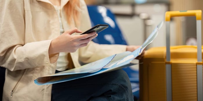 A young woman at the airport with luggage and a map, using her smartphone for travel planning and navigation. Ideal for themes of modern travel, solo female travelers, and travel preparation.