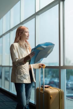 A woman stands in an airport terminal holding a map and suitcase, ready for her travel adventure. She looks out the window, planning her journey.