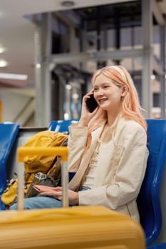 Young woman at airport, smiling and talking on phone, sitting with luggage, ready for travel. Modern travel concept.