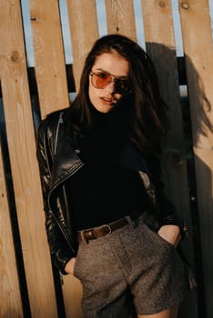 portrait of a stylish young brunette girl in sunglasses and a leather jacket on the background of a wooden fence in autumn