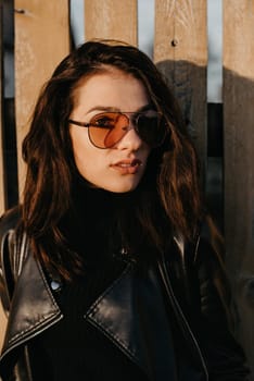 portrait of a stylish young brunette girl in sunglasses and a leather jacket on the background of a wooden fence in autumn